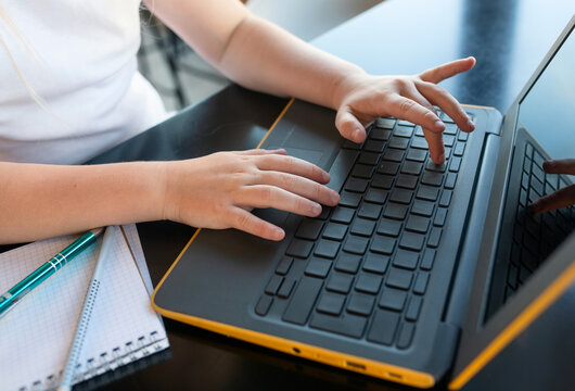 A child is typing on a laptop at a black desk. There are notebooks and pens nearby. The scene suggests studying or remote learning in a bright, modern indoor setting.