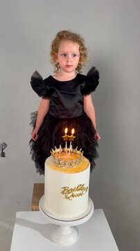 Little girl in black dress standing behind birthday cake with topper. Child celebrating birthday, wearing crown. Festive party decoration and dessert concept.
