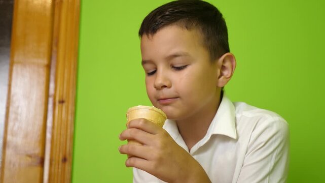 Boy in a white shirt eats an ice cream cone, taking a bite of a sweet frozen dessert against a green background.