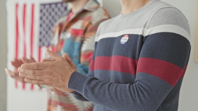 Two men clapping hands with an i voted sticker on sweater and a visible american flag on wall in a polling building; civic pride.