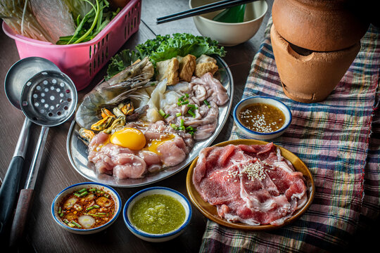 Traditional Thai clay pot hot pot known as Jim Jum, featuring raw sliced pork, fresh seafood, egg, vegetables, and spicy dipping sauces on a wooden table, perfect for an Asian food concept. 