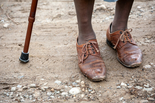 Close-up of worn leather shoes and a walking cane on a muddy rural path, symbolizing poverty, disability, aging, resilience, mobility and everyday hardship.