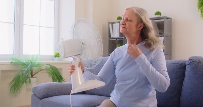 Senior elderly woman sitting on sofa in living room at home suffering from summer heat in front of fan. Female retired person using electric fan enjoying cool air during heatwave. 4k video.