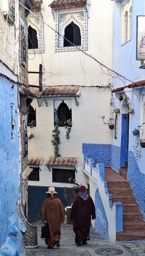 Two local women in traditional djellabas walking through a picturesque narrow street with Arabic architecture in Chefchaouen.