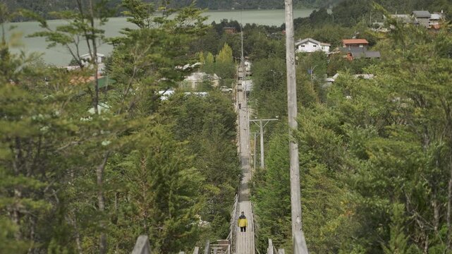 Elevated view of a person in a yellow jacket walking along a long wooden boardwalk, leading towards the rural coastal village of Caleta Tortel, Chile.