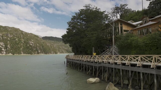 Person in a yellow jacket walks along a wooden boardwalk beside a house, overlooking a bay in the remote coastal village of Caleta Tortel, Chile.