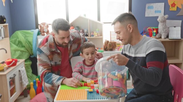 Man and boy assemble colorful plastic blocks, one man pours bricks from a bucket while the other guides child's hands at a play table inside a building; playful bonding joy.