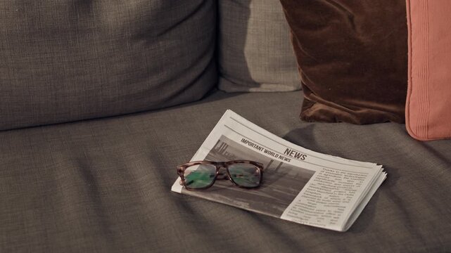 Close up of newspaper and eyeglasses resting on grey sofa, illustrating morning routine and staying informed. Footage suitable for media, journalism, retirement, and lifestyle marketing campaigns