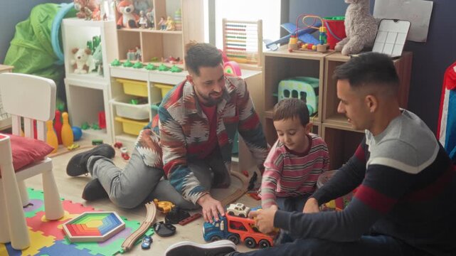 Boy reaches hand for toy car while two men guide play and hand blocks on foam mat in cluttered playroom; family bonding joy.
