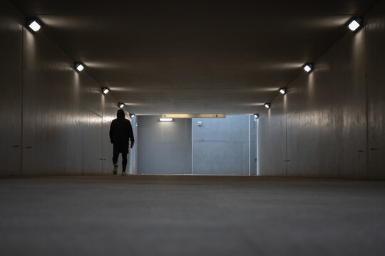 Silhouette of a man in dark hoodie walking away inside an empty concrete pedestrian underpass tunnel illuminated by symmetrical rows of square lamps toward city street exit transit area.
