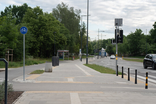 Urban pedestrian sidewalk with yellow tactile paving features a shared bicycle path sign traffic lights a commuter bus stop shelter and cars on asphalt road with green trees background area.