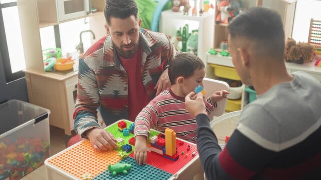 Boy reaches with hand for plastic block on colorful play table while two men assist in building; nurturing learning bonding joy.
