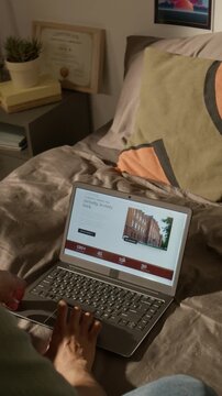 Vertical video of hand of unrecognizable young student lying in bed, scrolling through college website on laptop. Clip is ideal for university enrollment ads and higher education promotional videos