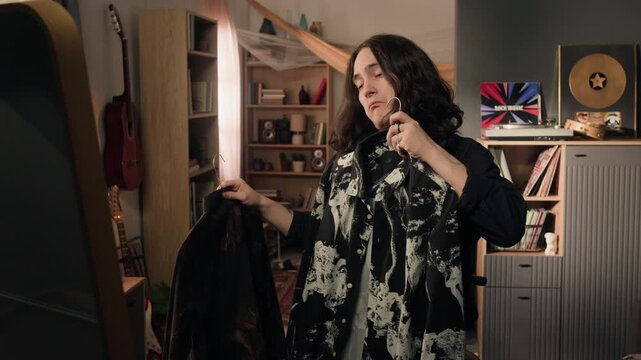 Young man with long hair standing in front of mirror, holding two patterned shirts on hangers and choosing outfit. Perfect for fashion retail and lifestyle marketing campaigns