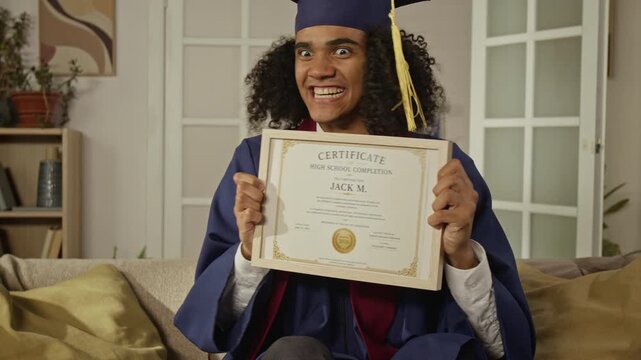 Tilt up portrait of happy African American male teenager wearing graduation gown, showing high school completion certificate to camera, celebrating online graduation. Perfect for e-learning ads