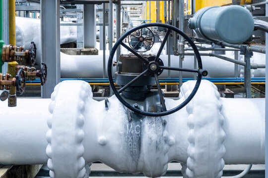 Close-up of a large manual gate valve on frozen pipelines covered in ice due to cryogenic gas flow inside an LNG or industrial plant surrounded by yellow-painted steel structures.
