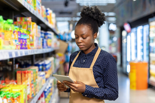 Woman employee wearing an apron and working in a supermarket aisle, checking product stock and managing inventory with a modern digital tablet