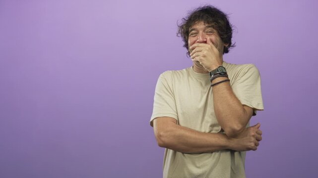 Young hispanic man with hand covering mouth and crossed arm, wearing beige tshirt and wristwatch, posed against a purple studio backdrop; shy amusement.