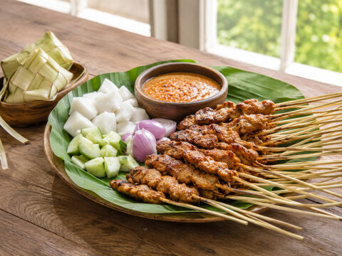  Brochetas de carne satay a la parrilla servidas sobre una hoja verde con salsa de cacahuete, pepino, cebolla y dados de arroz, presentadas sobre una mesa de madera junto a paquetes tradicionales de h