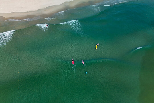 People paddleboard and surf on clear, blue-green ocean water. A sandy beach curves along the shore, with small waves breaking closer to land