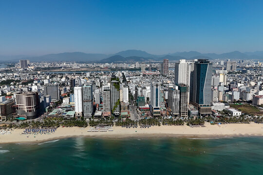 Panoramic view shows a bustling coastal city shoreline. A wide sandy beach is filled with umbrellas and people