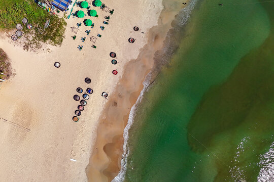 People relax under green umbrellas on a sandy beach. Numerous traditional basket boats rest on the shore and near the gentle waves of the clear ocean