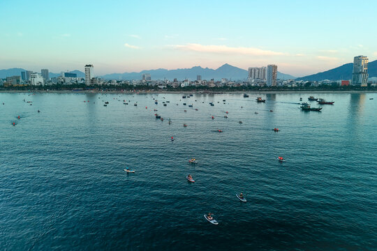 Numerous stand up paddleboarders and boats navigate the calm waters of Da Nang Bay, Vietnam. A city skyline and mountains rise under a soft morning sky