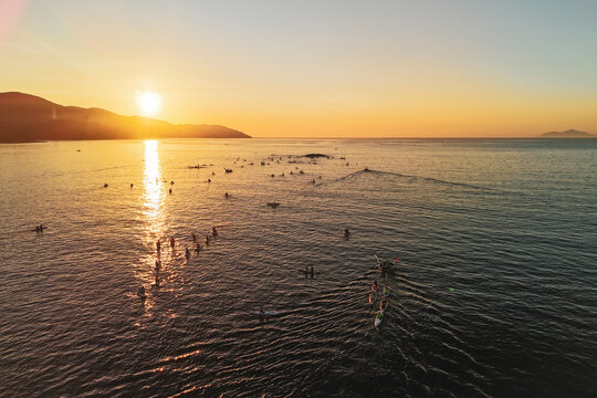 Numerous paddleboarders and small boats navigate calm ocean waters during a vibrant golden sunset. Mountains line the horizon with a distant island visible
