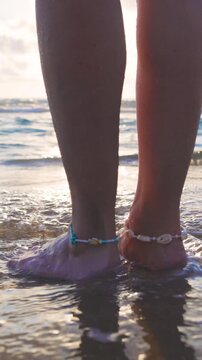 vertical video. A close-up of bare lower legs and feet standing on wet sand at the waters edge. anklets are visible, and the scene is bathed in the warm light of sunset. Summer mood.
