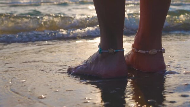 A close-up of bare lower legs and feet standing on wet sand at the waters edge. anklets are visible, and the scene is bathed in the warm light of sunset. Summer mood.