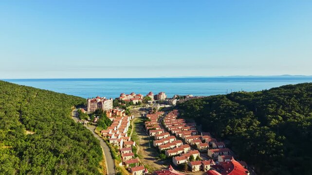 Elenite is rebuilding after a flood that impacted this coastal town. The landscape shows affected housing and greenery under a clear sky. The sea is visible in the distance.