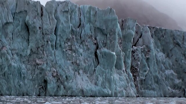 Witness the breathtaking spectacle of an ancient Arctic glacier dramatically calving. Chunks of ice plummet into the frigid ocean below a towering, sculpted ice wall on a misty day.
