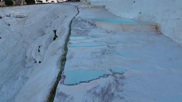 A drone pulls back and ascends from the natural thermal water, revealing a wide scenic view of Pamukkale's white travertine terraces. Soft dawn light reflects on the pools as the landscape opens up un