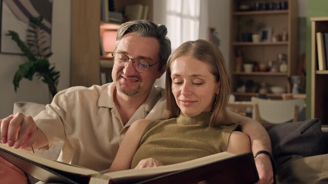 Close up of middle aged Caucasian couple sitting on sofa and looking through photo album together. Perfect for family heritage, memory preservation, or photo book printing service commercials