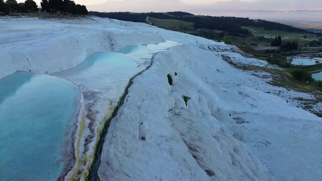 A smooth drone pan movement shifts from the white travertine terraces down to the scenic natural lake below Pamukkale. Soft dawn light reflects on the thermal pools and the water surface, framing a qu