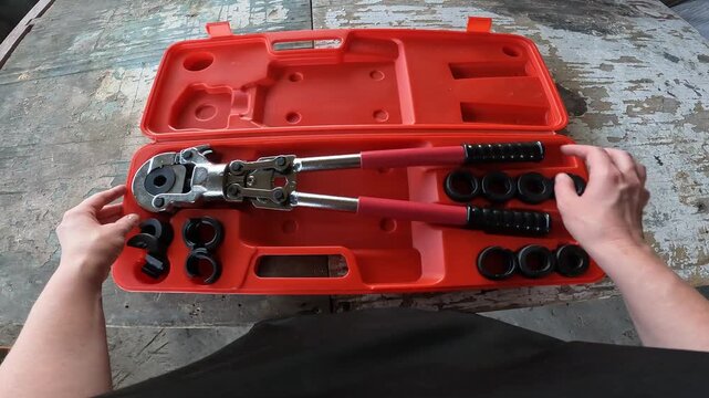 Manual crimping pliers for metal-plastic pipes in a case. Plumber holds a professional crimping tool for installing PEX pipes. Top view of a case with interchangeable nozzles on a wooden background