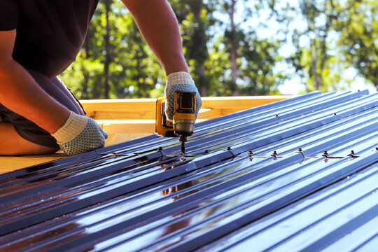 Roofing contractor uses power tool to attach metal panels on roof at building site.