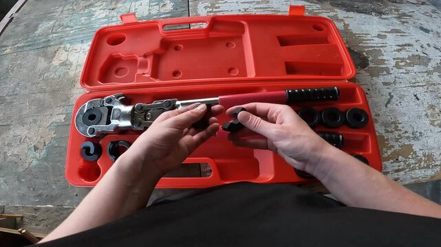 Manual crimping pliers for metal-plastic pipes in a case. Plumber holds a professional crimping tool for installing PEX pipes. Top view of a case with interchangeable nozzles on a wooden background