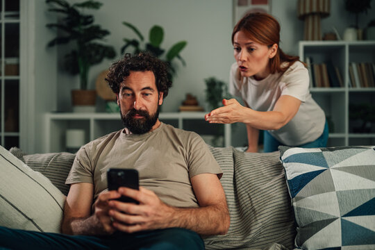 Man absorbed in smartphone ignoring angry woman arguing in background, representing relationship problems and digital addiction