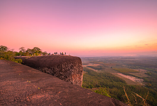 Epic sunrise at a famous Thai viewpoint, capturing tiny human figures on a colossal rock ledge above a sea of trees, evoking adventure, scale, and serenity.