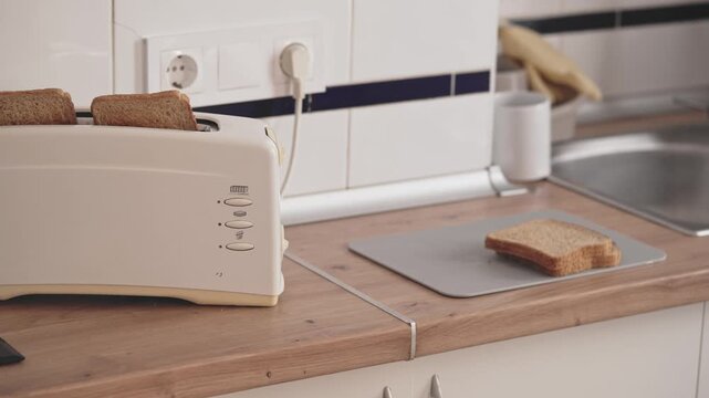 In a kitchen, a toaster warms slices of bread on a counter. The kitchen has a sink and other kitchen tools. The setting is bright in the morning hours.