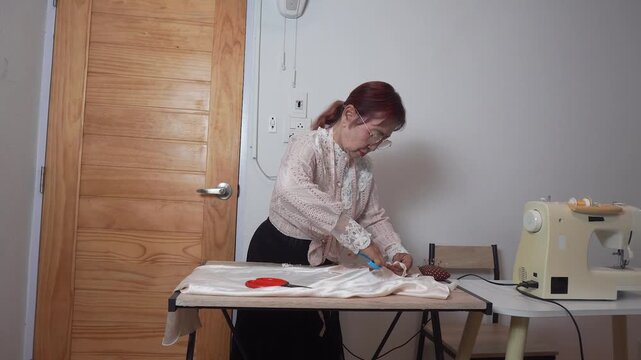 Elderly woman with measuring tape around neck uses scissors to cut clothes at home. Senior tailor works her creative hobby during retirement lifestyle.