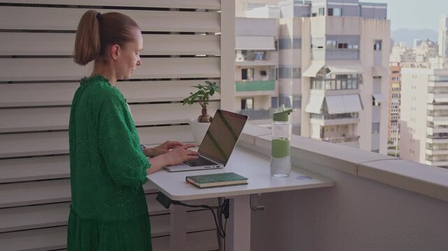 A person sits at a small table on a balcony. They work on a laptop while looking out at the skyline of the city. The sun shines brightly and the view is clear.
