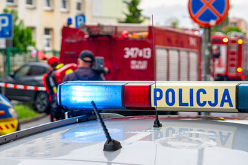 Close-up of a police car light bar with the text "POLICJA" in the foreground, with a blurred background showing an accident scene with red fire trucks, firefighters, and emergency response operations, © Adam