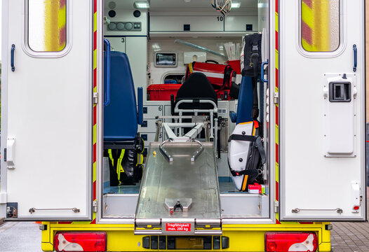 Interior of a modern emergency ambulance viewed through the open rear doors, featuring a metal stretcher ramp with a "Tragf&auml;higkeit max. 280 kg" sticker, medical seats, rescue backpacks, bags, and equ