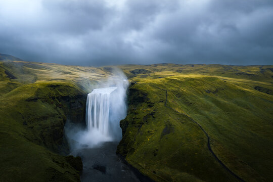 Aerial view of Skogafoss waterfall cascading down lush green cliffs under a dramatic cloudy sky in Vik, Myrdalshreppur, Iceland.