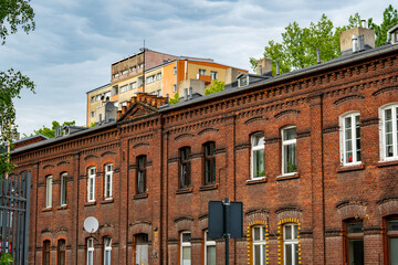 Historic red brick tenement house building typical for Lodz architecture with a modern apartment block in the background under a cloudy sky in daylight. © Adam