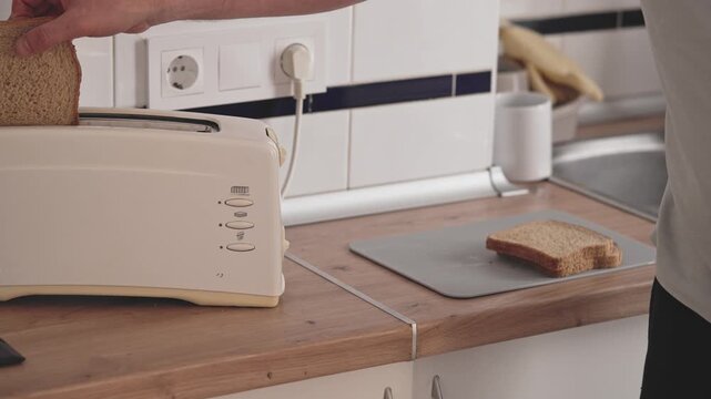 A person places slices of bread into a toaster on a kitchen counter. They then press the lever down to start toasting. This often happens during breakfast preparation.