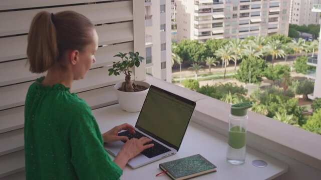 A person is typing on a laptop while sitting on a balcony. The sun is shining and there are green plants nearby. The city is visible in the background. This is a nice space for work.