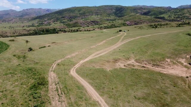 Wide angle drone shot showcasing a network of dirt roads on a dry grassy plateau, with a tiny white car in the distance and rugged mountain ridges in Kazakhstan.
